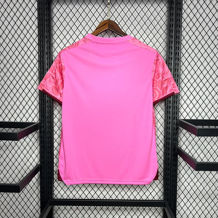 Maillot de football rose avec motifs sur les manches, suspendu sur un cintre devant un mur en treillis métallique