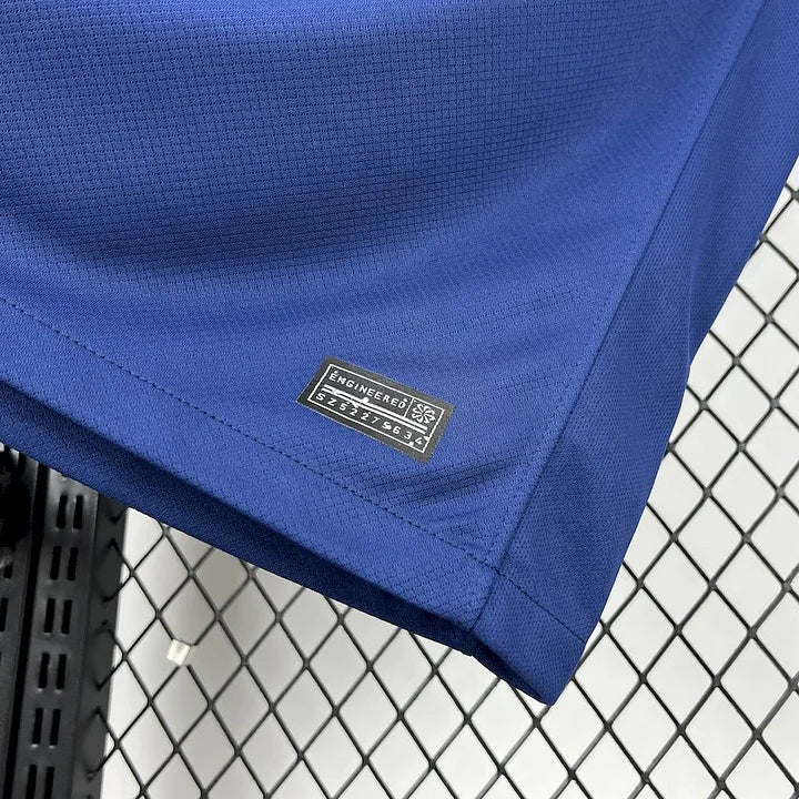 Détail du tissu bleu et étiquette "Engineered" sur maillot de football PSG 2025-2026 devant grille métallique